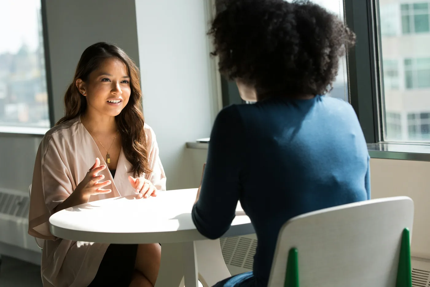 Two people talking across a table beside an office window.
