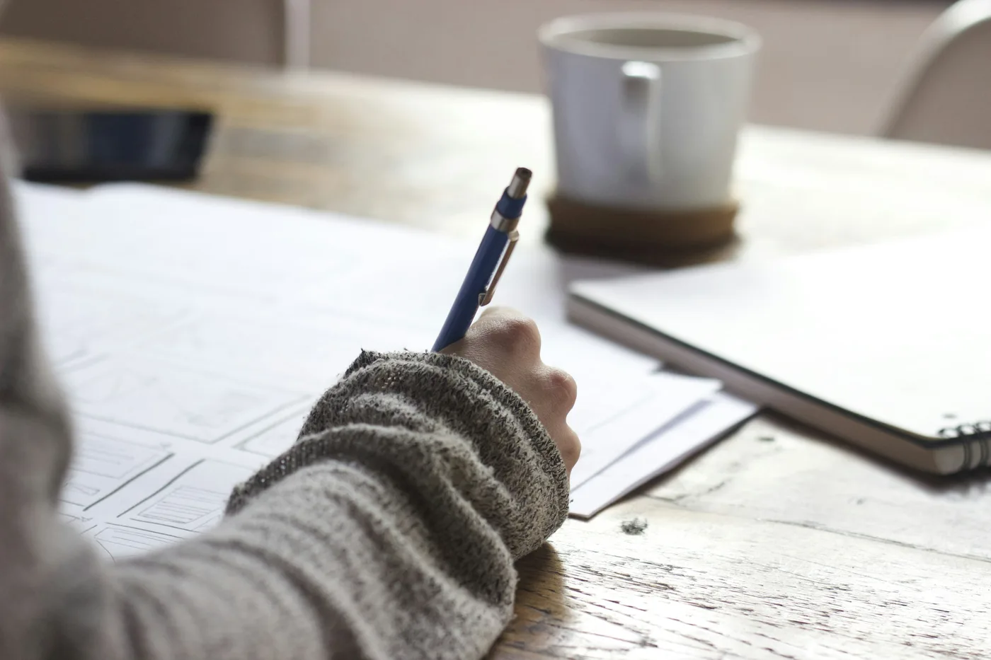A hand writing notes over technical drawings beside a laptop.